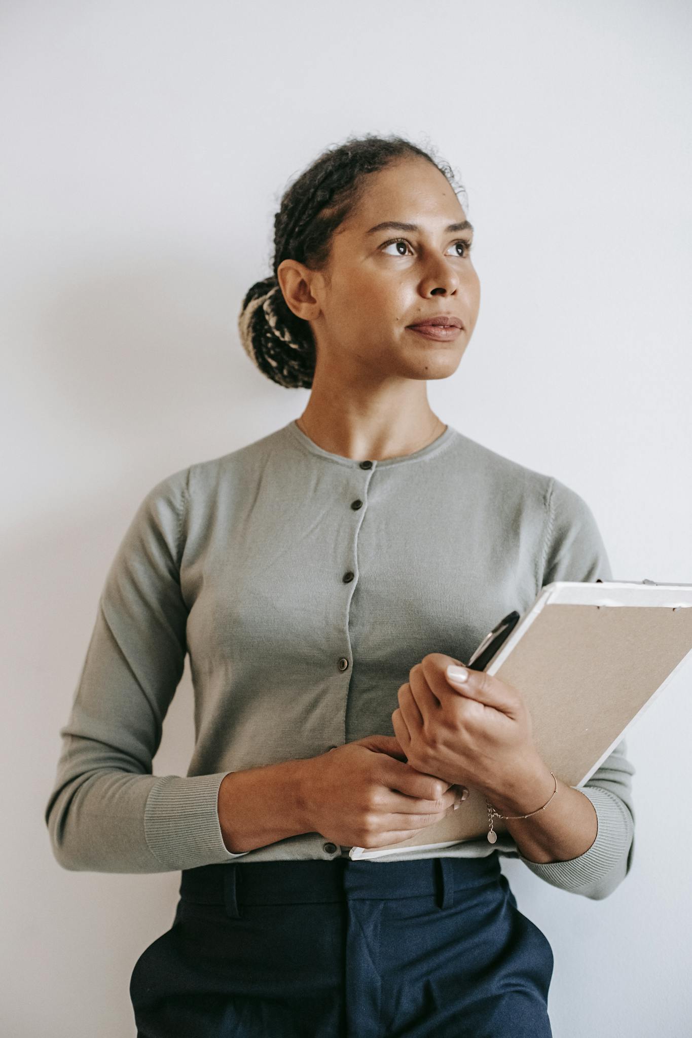 Hispanic woman with dark hair stands confidently indoors holding a clipboard, looking thoughtfully ahead.