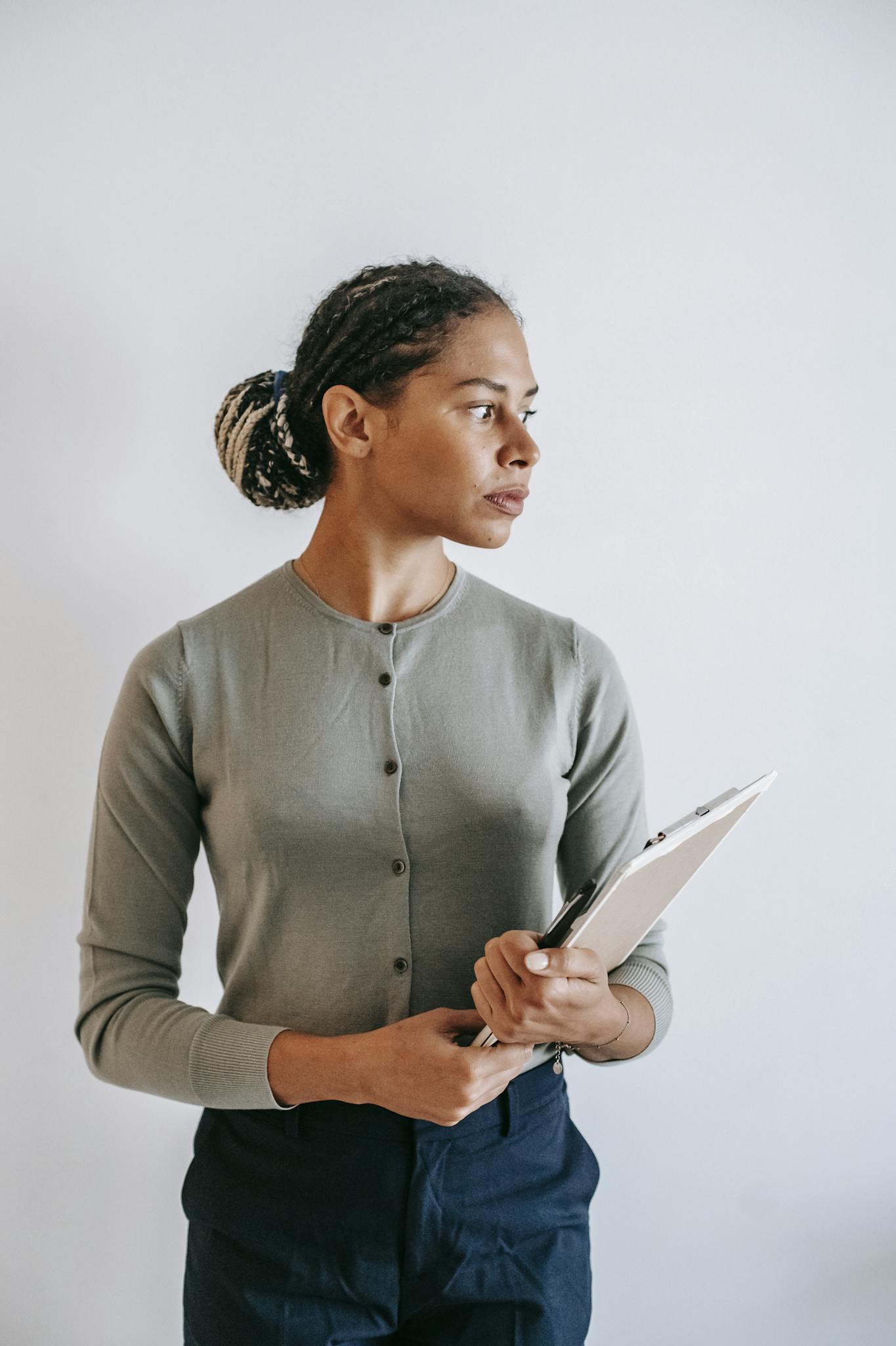 Serious ethnic female in formal wear with clipboard in hands standing against white wall and looking away in contemplation
